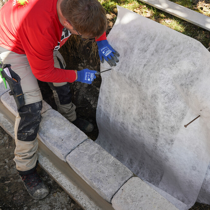 Heimwerker sichert das Drainagevlies mit langen Nägeln in der Baugrube im Garten.