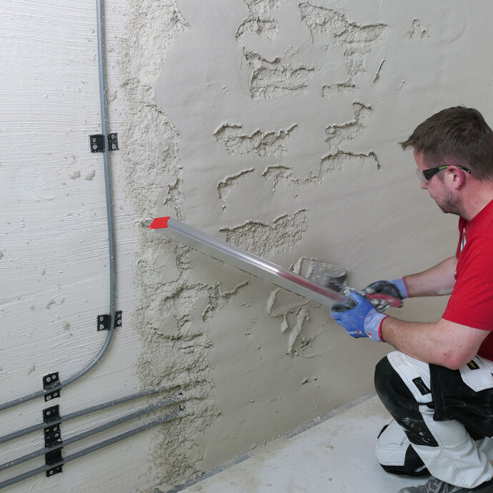 Mann bessert ungleichmäßig verputzte Stellen an der Wand mit Putzmaterial und Abziehlatte aus.