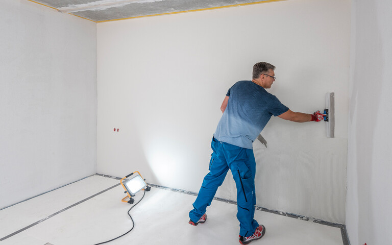 Heimwerker zieht im Raum einen weißen, feinen Oberputz mit einem Flächenspachtel ab. Um Unebenheiten zu sehen, wird die Wand von einem Baustrahler angestrahlt.