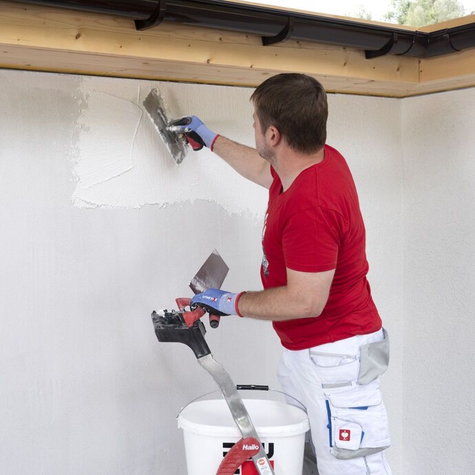 Mann steht auf Leiter und trägt Rillenputz von oben nach unten mit einer Glättkelle an der Wand auf.