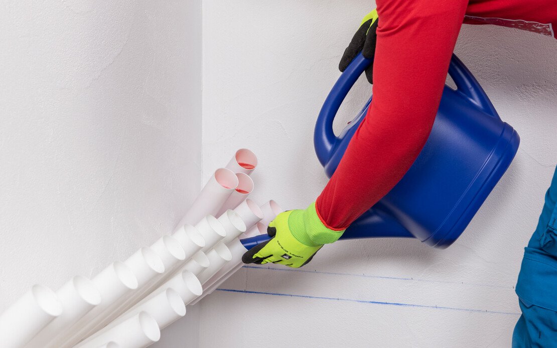 Person mit Schutzhandschuhen befüllt mit einer blauen Gießkanne Injektionstrichter in einer Wand zur Horizontalabdichtung.