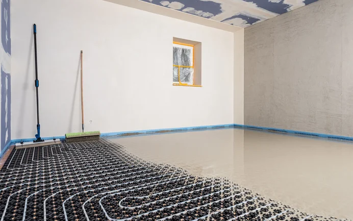 Foto eines Innenraumes: Bodenheizung ist verlegt, Schicht Ausgleichsmasse wird eingearbeitet. In der rechten Ecke stehen Werkzeuge - eine Spachtel und eine Stachelwalze.