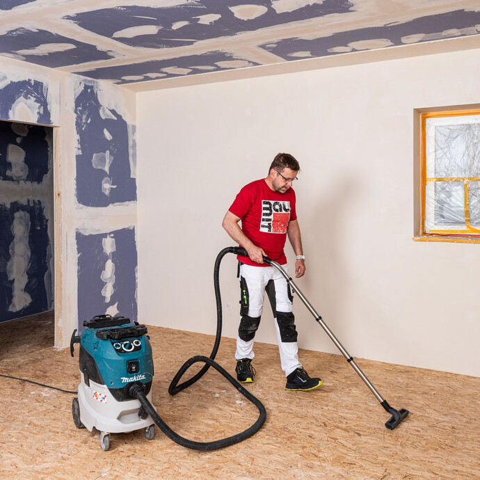 Un bricoleur portant un T-shirt rouge Baumit aspire le sol en bois avec un aspirateur industriel.