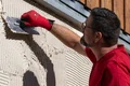 Un bricoleur applique un enduit extérieur sur une façade. Gant rouge Pacifique pour une prise en main sûre. Idéal pour les projets de bricolage.