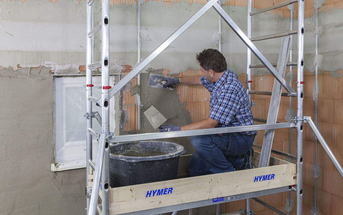 Mann tr&auml;gt Kalkzementputz auf den vorbereiteten Untergrund auf. W&auml;hrenddessen steht er auf einem Ger&uuml;st.