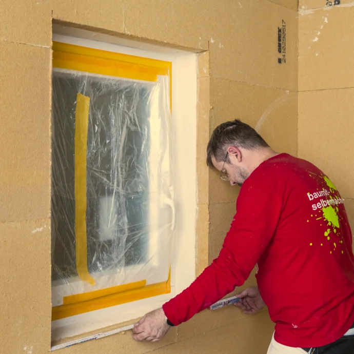 Bricolateur en train de masquer une fenêtre en vue de travaux de peinture, cadre de fenêtre préparé avec un film de protection et du ruban adhésif.