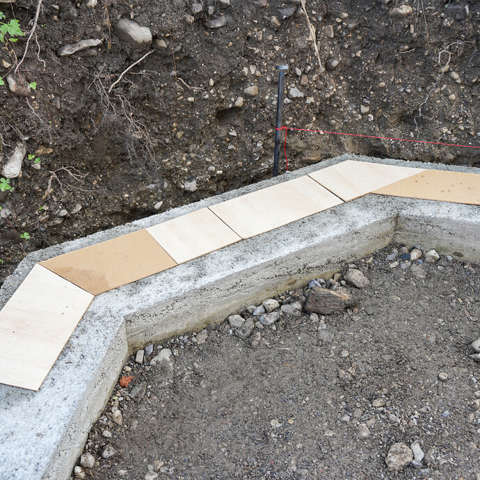 Selbst angefertigte Schablonen für die Mauersteine aus MDF-Platten liegen auf dem Fundament, um die Mauersteine richtig zuzusägen. Eine rote Richtschnur, die am Erdnagel befestigt ist, ist über dem Fundament gespannt.