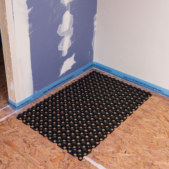 Rénovation d'un bâtiment ancien : pièce à rénover avec une cloison sèche et une bande d'isolation périphérique fixée. La bande à excroissances Minitec est positionnée sur le plancher en bois dans le coin de la pièce.