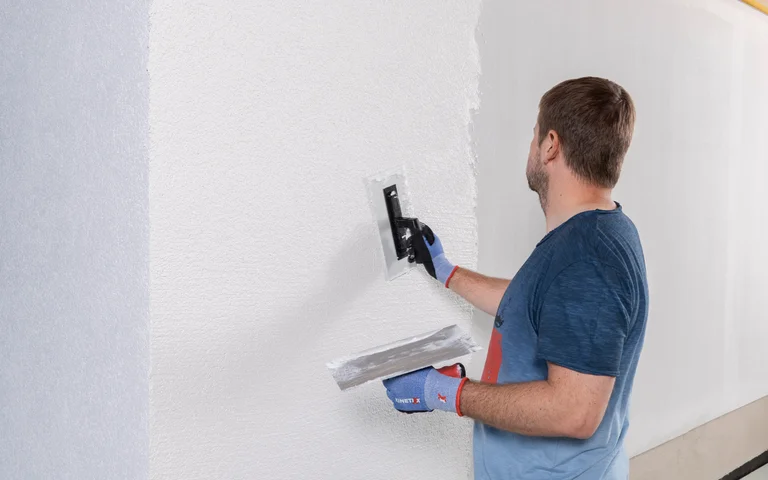 Un bricoleur enduit un mur intérieur à l’aide d’une truelle, en appliquant de l’enduit blanc à l’horizontale, une source d’inspiration pour les projets de bricolage.