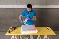 Mann trägt Kleber auf Dämmplatte auf, Heimwerkerprojekt mit Baumit-Produkten auf gelbem Arbeitstisch im Innenbereich.