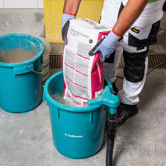 Detailfoto: Person schüttet die Ausgleichsmasse Extrem von Baumit mit Handschuhen in einen blauen Eimer von Collomix.