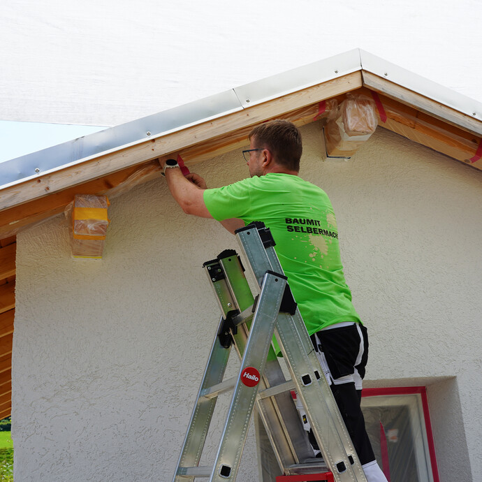 Mann steht auf einer Leiter und klebt Sparren und Holzbretter am Dach mit Folie und Klebeband ab.