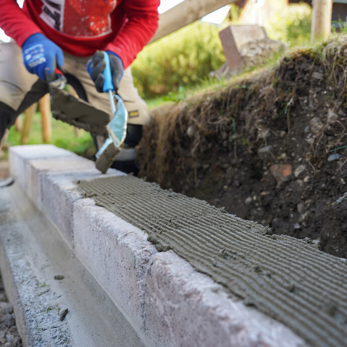 Steinkleber wird mit einer Klebekelle gleichmäßig auf eine Mauer aus Betonsteinen aufgetragen.