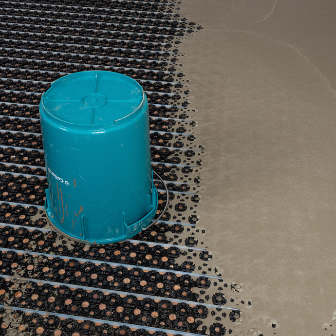 Le seau bleu vide est posé à l'envers sur le chauffage au sol nouvellement installé avec une plaque à plots. A droite, on peut voir la masse d'égalisation fraîchement vidée.