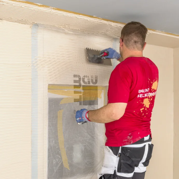 Heimwerker trägt Putz auf Wand auf; verwendet Zahnspachtel für gleichmäßige Verteilung. Schritt für Schritt Renovierung.