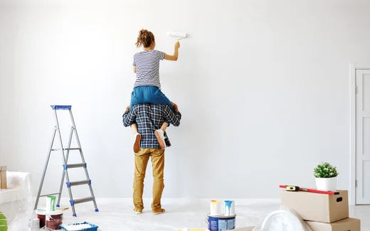 Paar beim Streichen einer Wand, Frau auf Leiter, Baumaterialien im Vordergrund, DIY-Heimwerkerprojekt.