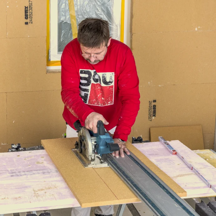Un bricoleur découpe un panneau de bois à l'aide d'une scie circulaire sur un établi, dans le cadre d'une rénovation ou d'un aménagement.