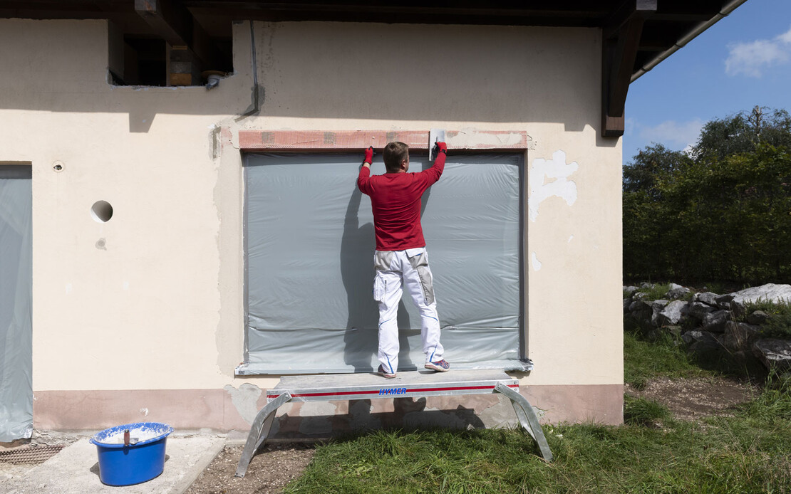 Heimwerker steht auf einer Leiter und setzt und verputzt den oberen Gewebeeckwinkel ein der Laibung der Terrassentüre.