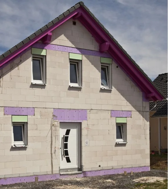 Maison individuelle non enduite, avec des avant-toits et des encadrements de fenêtres rehaussés de violet, avant l’enduction et l’isolation.