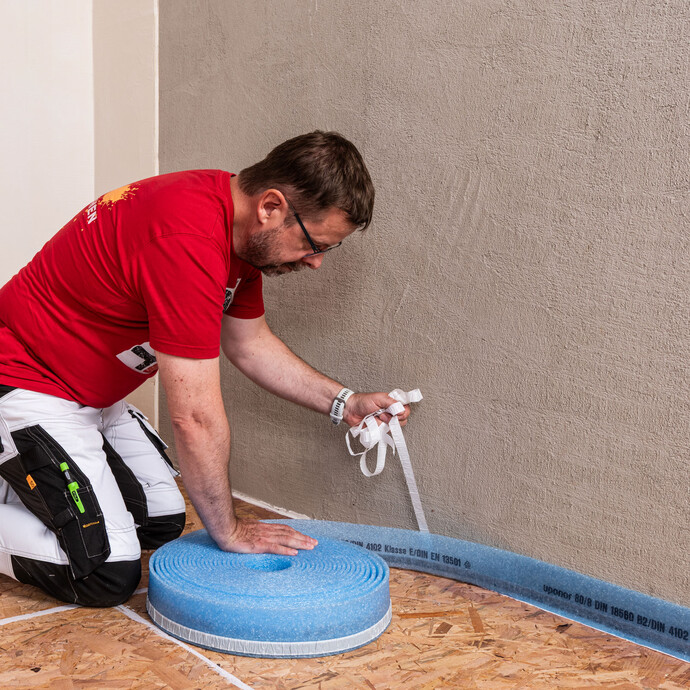 Le bricoleur fixe la bande d'isolation périphérique sur le mur. Il retire parallèlement le film de la bande isolante autocollante.