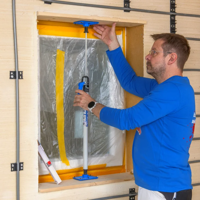 Heimwerker richtet Fensterrahmen mit Montagekeil aus, um Isolierung anzubringen. Fassade mit Dämmplatten verkleidet.
