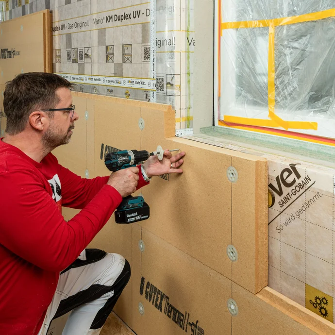 Baumit Handwerker verschraubt mit einem Schraubdübel die Holzfaserdämmplatte im Kantholz am Fenster. Innendämmung mit Tragkonstruktion zum Selbermachen.