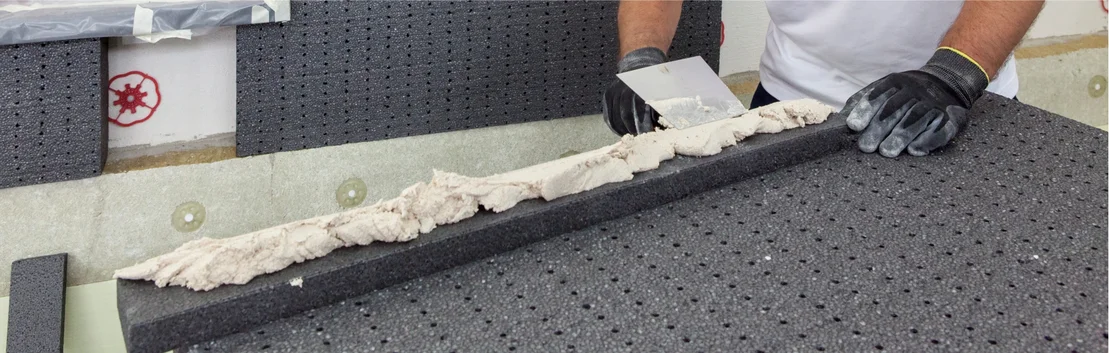 Un bricoleur applique de la colle sur un panneau isolant gris ; pr&eacute;paration en vue de la pose au mur.