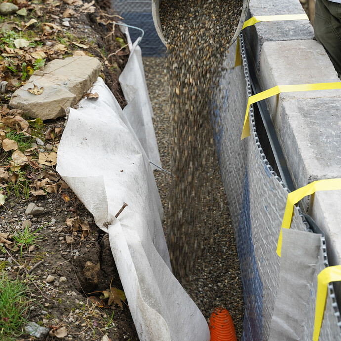 Detailaufnahme: Eimer mit Kies wird in die Baugrube auf das verlegte Drainagerohr gelegt.