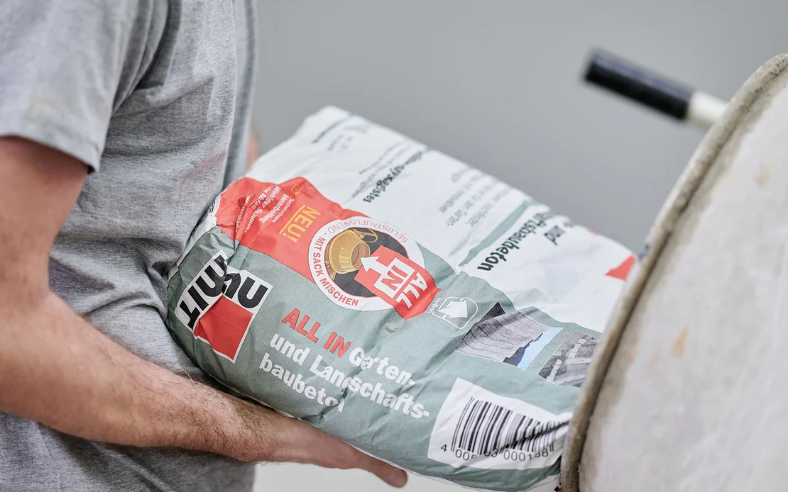 Person mischt Baumit Zementmörtel für Heimwerkerprojekte, hält Sack neben Betonmischer bereit. Ideal für Garten- und Landschaftsbau.