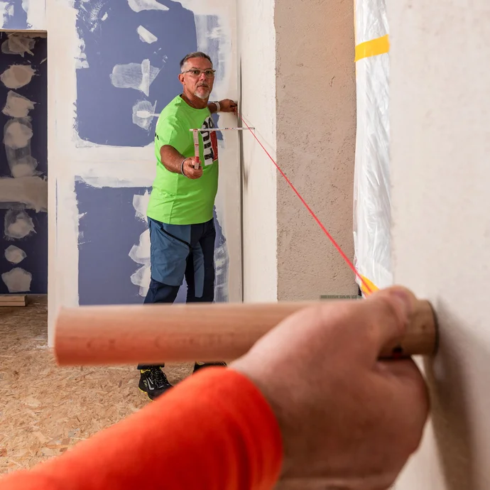 Zwei Heimwerker spannen eine Maurerschnur, um Unebenheiten an der Wand, wie z. B. Wellen oder Dellen, zu finden. Mit dem Meterstab wird der Abstand zur Wand entlang der Schnur gemessen.