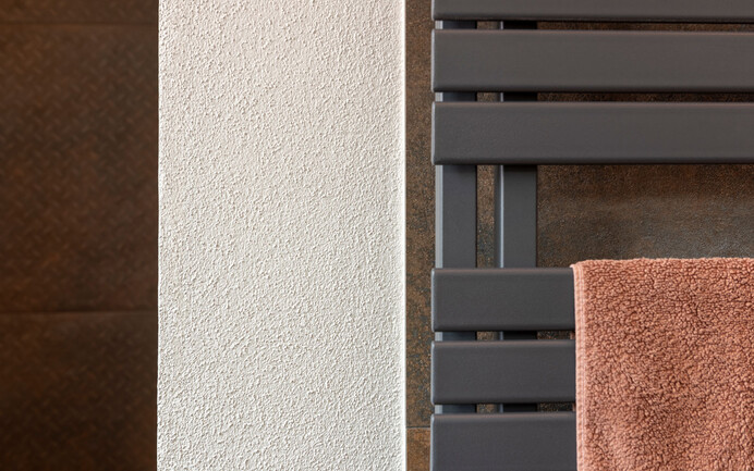 Détail de design moderne dans la salle de bains avec différents panneaux et porte-serviettes sombres de couleur rouille parfaitement mis en scène avec les murs blancs brillants à la fine structure feutrée avec l'enduit noble Baumit InStyle Edelweiss.