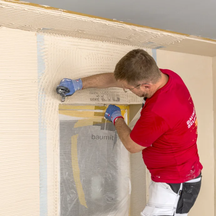 Heimwerker trägt mit Spachtel eine Putzschicht auf eine Innenwand auf, mit Baustoffen von Baumit.