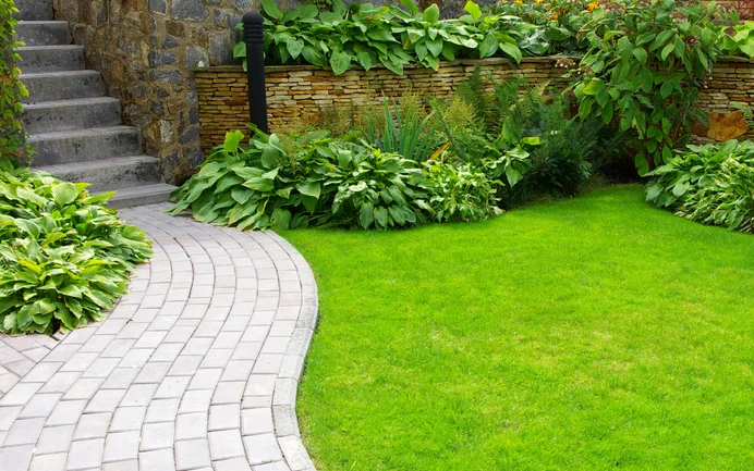 Gepflegter Gartenweg aus Natursteinpflaster führt entlang grünendem Rasen und üppiger Gartenbepflanzung zur Steintreppe.