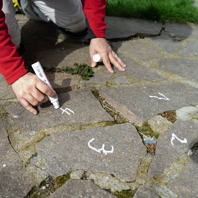 Person markiert lose Natursteinplatten mit einem weißen Kreidestift zur späteren Wiederverlegung.“