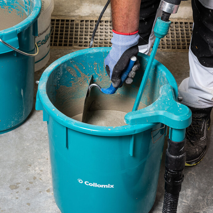 Nahaufnahme Foto: Blauer Eimer mit angerührter Ausgleichsmasse. Person streift mit einer Kelle die Mörtelreste am Rand des Eimers ab.