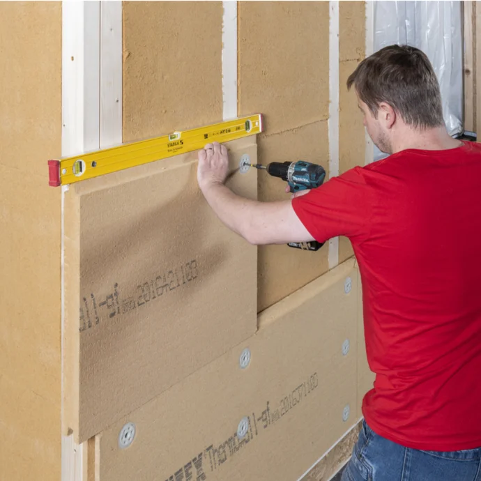 Heimwerker befestigt Dämmplatte an der Wand mit Bohrmaschine, Wasserwaage für präzises Arbeiten verwendend.
