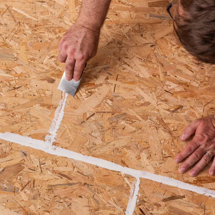 personne retire à l'aide d'une spatule la colle de montage appliquée, qui a été appliquée dans les joints des panneaux d'aggloméré, en affleurant la surface.