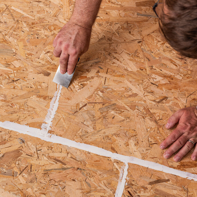 personne retire à l'aide d'une spatule la colle de montage appliquée, qui a été appliquée dans les joints des panneaux d'aggloméré, en affleurant la surface.