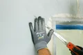 Heimwerker glättet Wand mit Spachtel neben Fensteröffnung, trägt Arbeitshandschuhe.