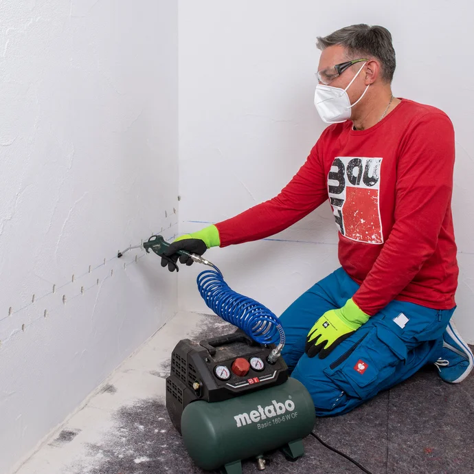 Handwerker entfernt mithilfe einer Handpumpe Staub aus dem Bohrloch. Zum Schutz trägt die Person eine Schutzbrille sowie einen Mundschutz und Arbeitshandschuhe.