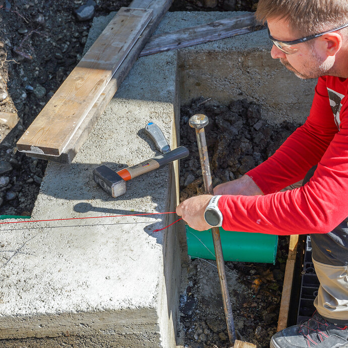 Eine Person mit Schutzbrille spannt eine rote Richtschnur am Erdnagel über dem Fundament, um die Mauersteine der Gartenmauer fluchtgerecht auszurichten. Ein Hammer liegt auf dem Fundament.