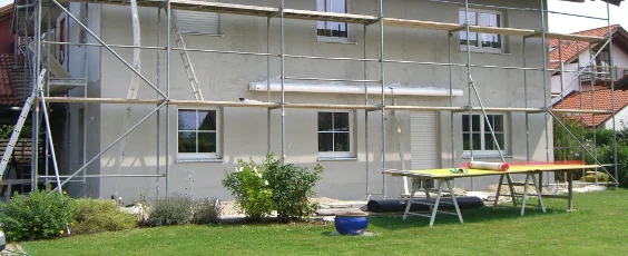 Façade de maison avec échafaudage pour des travaux de rénovation, vue de face avec surface préparée pour la peinture de façade, jardin verdoyant au premier plan.