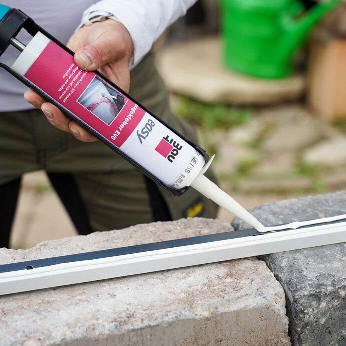 Handwerker trägt mit einer Kartuschenpistole Montagekleber entlang eines Abschlussprofils auf einer Mauerkrone auf.