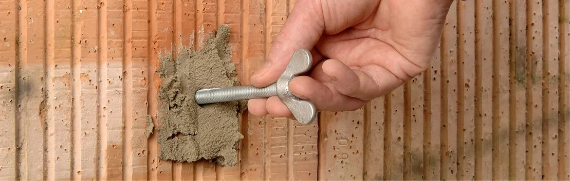 Hand zieht Metallschraube aus einer mit M&ouml;rtel gef&uuml;llten Klinkerwand, zeigt Reparaturstellen-Vorbereitung.