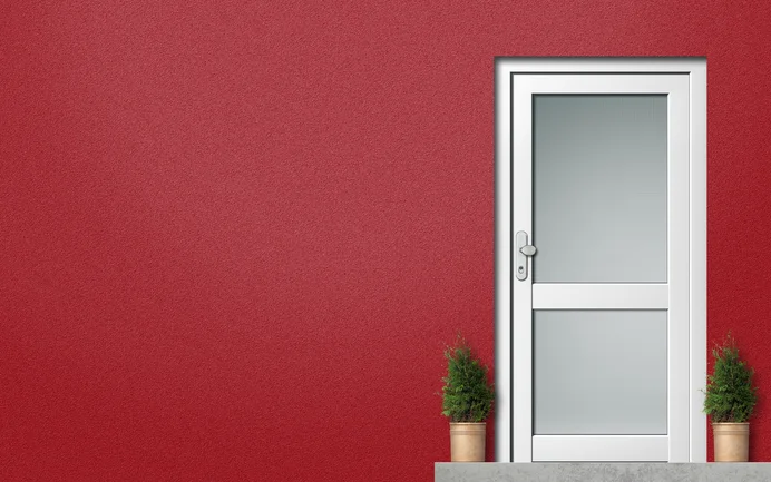 Porte d’entrée blanche sur une façade en crépi rouge, flanquée de deux petites plantes en pot.