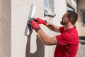 Un homme enduit un mur extérieur à l’aide d’une taloche de lissage, il porte des gants rouges, à la lumière du jour.