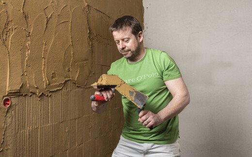 Person trägt mit Hilfe einer Kelle den Lehmputz an der Wand im Innenbereich auf.