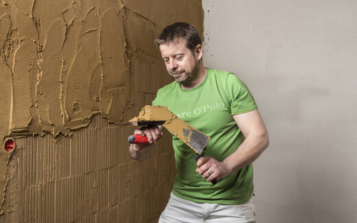 Person trägt mit Hilfe einer Kelle den Lehmputz an der Wand im Innenbereich auf.