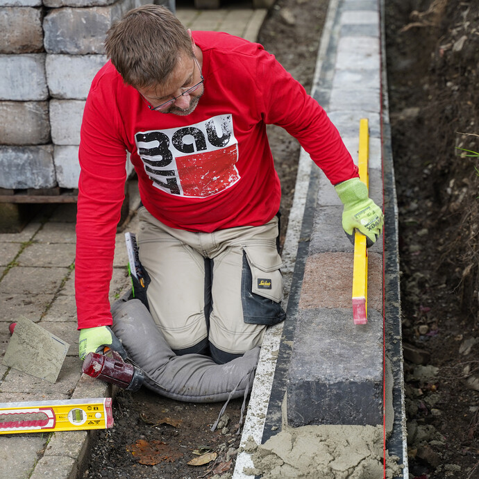Eine Person kniet auf dem Boden und prüft mit einer langen Wasserwaage die Lage gesetzter Mauersteine. Im Hintergrund liegen Steinpakete bereit, neben der Mauer ist frischer Mörtel zu sehen.