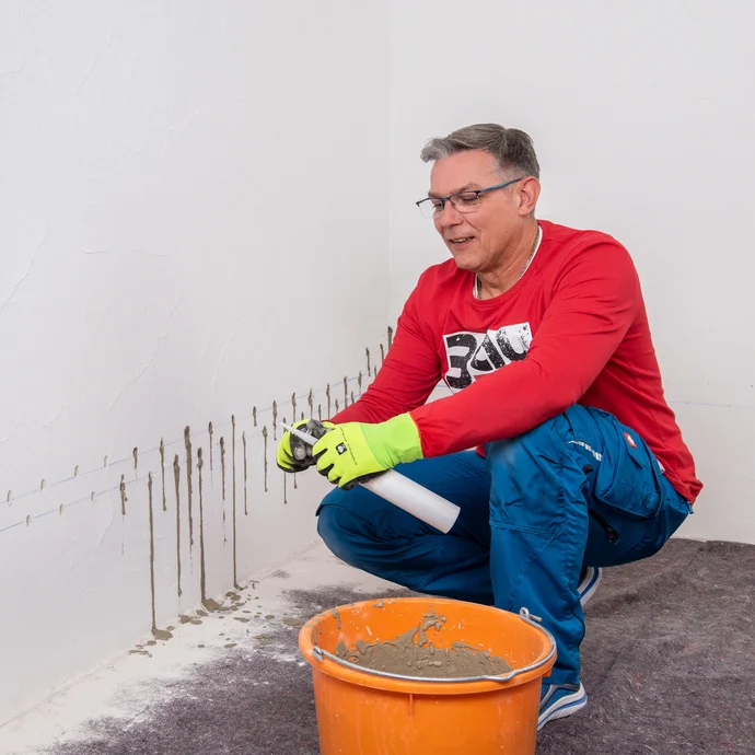 Handwerker bereitet einen weiteren Injektionstrichter vor. Die Einfüllspitze wird mit einem Cuttermesser gekürzt. In einem Eimer ist der bereits angemischter Betonspachtel.